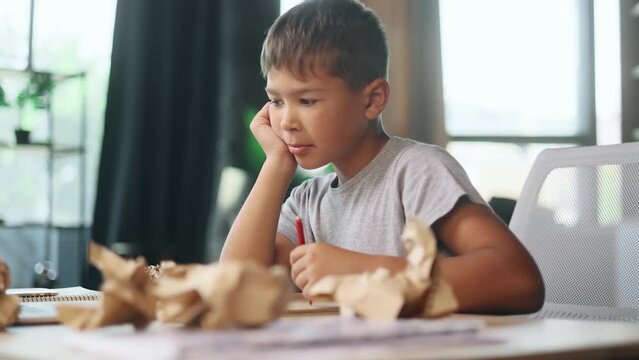 Portrait of exhausted school boy with failed crumped papers while writing in notebook at home Tired little kid child with creative block or lack of inspiration indoors Stress education concept
