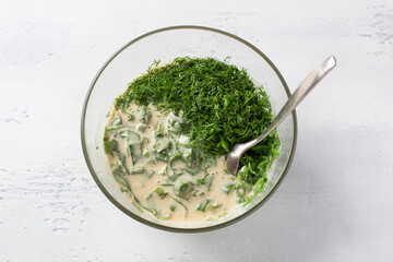 Glass bowl with spinach and herb pie dough, spinach, green onion and dill added to the egg-yogurt mixture on a light blue background, top view. Dough kneading, cooking stage
