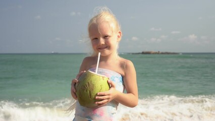 Child girl drinks coconut milk from a coconut on a tropical beach against the backdrop of the sea