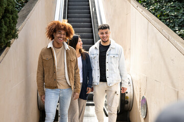 Diverse group of friends going down an outdoor escalator. Three happy young people smiling and looking relaxed walking down of stairs in the street.