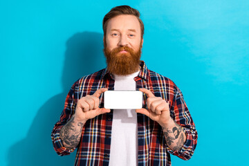 Portrait of friendly man with tattoo wear plaid shirt hold horizontally smartphone screen empty space isolated on blue color background