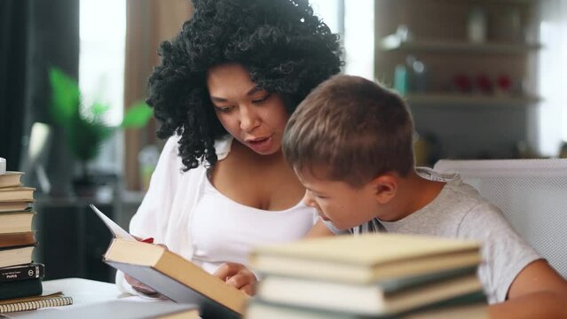 Cute primary school boy kid studying with his mother reading book at home Charming young woman mom helping to her son doing homework together indoors Distance online education