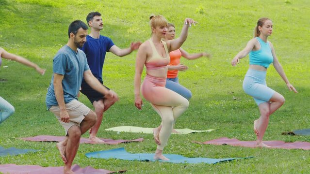 Female coach explaining and demonstrating eagle pose to diverse group of young people in sunny summer park. Yoga practitioners balancing on one leg and practicing together. Health and wellness concept