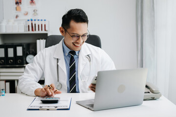Confident Young Asian male doctor in white medical uniform sit at desk working on computer. Smiling use laptop write in medical journal in clinic.