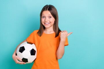 Portrait of schoolgirl with straight hair dressed orange clothes hold ball directing empty space isolated on turquoise color background