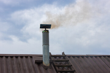 Roof of a house with a metal chimney with smoke coming out of it against the sky