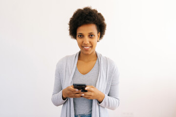 Studio portrait of happy African American young woman holding smartphone typing browsing looking at camera with friendly expression. Black female holding mobile cellphone on isolated white background.