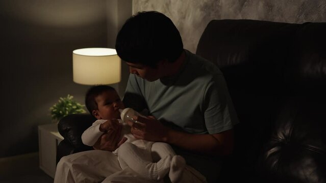 stressed father trying to feeding milk bottle to crying infant baby on a sofa in the living room at night