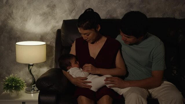 father and mother breastfeeding infant baby on a sofa in the living room at night