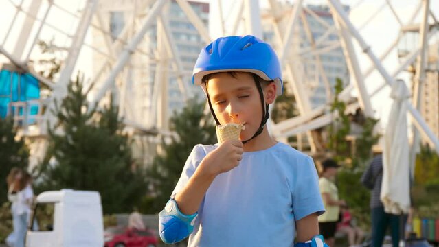 A happy child eats ice cream. A boy in a skateboard helmet. A child in a city park on the background of a Ferris wheel. Active recreation of children. street food. urban development