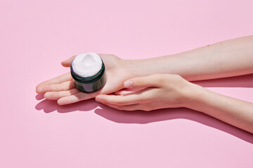 Cropped top view of female hands holding black jar of cream, night cream in hands. Using cosmetics product isolated pastel pink background.