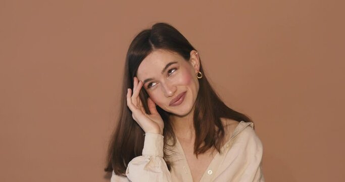 Shocked shy charming confused young woman in beige shirt posing isolated on beige background studio. Girl looking at camera smile hold hands on head, cover face.