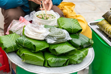 Close up view of a tray with banh tet or banh chung which is sticky rice cake with a person sitting next to the tray with a traditional bowl of soup or pho