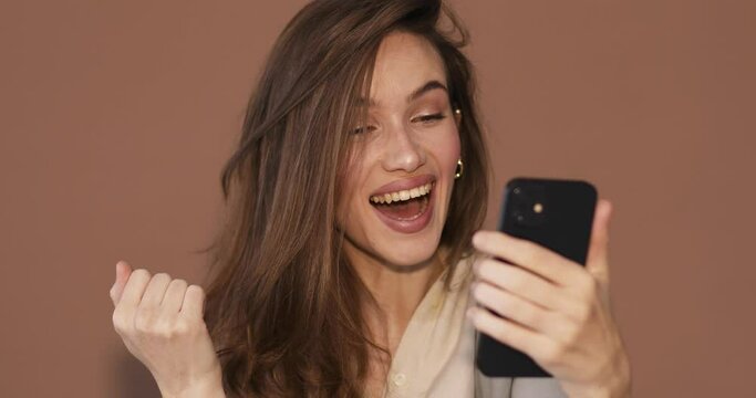 Attractive brunette woman look surprised wow hold using mobile cell phone on beige background. Girl look on smartphone and doing winner gesture. Girl just found out great big win news, sales, lottery.