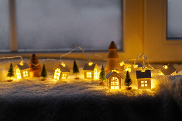 Atmospheric miniature winter village. Stylish cute little glowing houses and christmas trees on soft snowy blanket in evening room. Christmas hygge background. Happy Holidays!