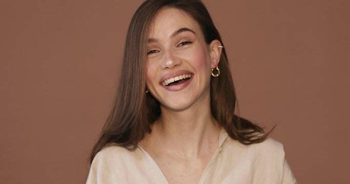 Friendly girl makes hello gesture glad to meet friend. Hi nice to meet you. Brunette girl wear beige shirt and gold earrings wave hand isolated on beige background. Girl look flirting and friendly.