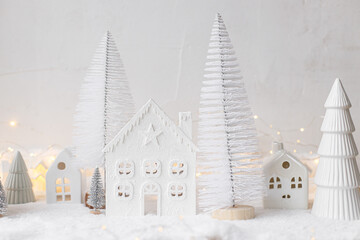 Cozy christmas miniature village. Stylish little ceramic houses and trees on snow blanket with golden lights on white background. Atmospheric winter village modern still life. Merry Christmas!