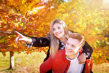 Happy autumn (fall) couple. Boyfriend carrying girlfriend on piggyback. Girl showing with...