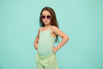 Emotional authentic confident strong kid girl in sunglasses, puts her hands on waist and looking confidently at camera