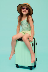 Full length vertical portrait of a Caucasian cute child girl 6 years old, sitting on her suitcase, isolated background