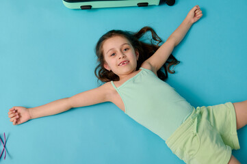 Mischievous child preschool girl with suitcase, lying on blue background, expressing positive emotions looking at camera