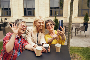 Smiling mature ladies sitting in cafe terrace and talking via video call with friend and waving hi