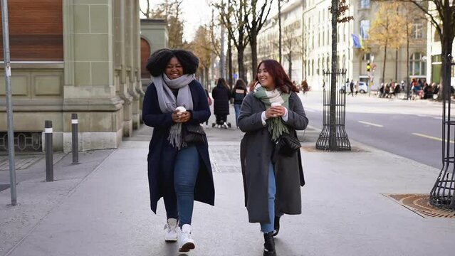 Young multiracial women drinking coffee to go and having fun walking together in the city during winter time