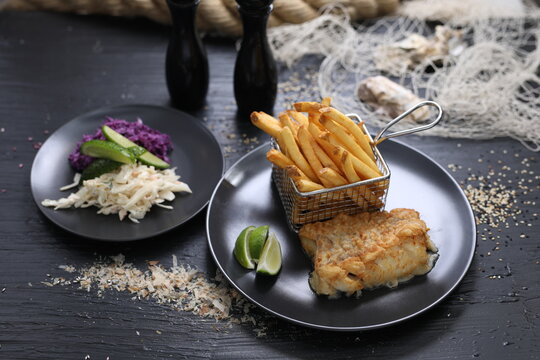 Fried fish fillets served with potato fries in a metal serving basket and salad mix, on black plates, selective focus.