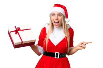 Young blonde woman with christmas hat and holding a gift over isolated chroma key background surprised and pointing finger to the side