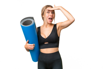 Blonde sport woman going to yoga classes while holding a mat over isolated chroma key background doing surprise gesture while looking to the side