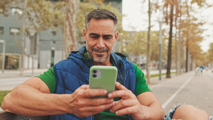 Fototapeta premium Close-up,happy mature man wearing casual clothes sitting on bench looking through information on smartphone