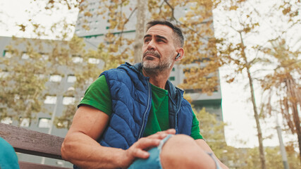 Mature man wearing casual clothes in wireless earphone enjoying music while sitting on park bench, bottom view, soft focus