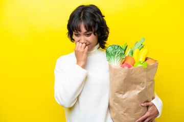 Young Argentinian woman holding a grocery shopping bag isolated on yellow background having doubts