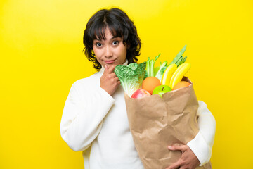 Young Argentinian woman holding a grocery shopping bag isolated on yellow background thinking