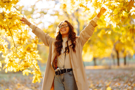 In the autumn park, a happy woman in stylish clothes and a camera is having fun among the fallen leaves. Smiling tourist woman enjoys warm sunny weather in autumn season.