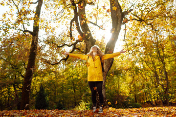 Happy female hiker in yellow coat walking in golden autumn forest. Beautiful woman has fun in...