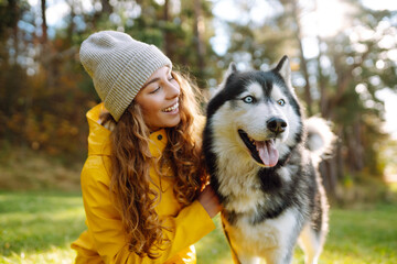 Beautiful young woman in a hat and yellow coat walks with her husky on the lawn in the park. Happy female posing outdoors with her dog. Concept of friendship, vacation, walk. © maxbelchenko