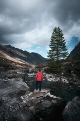 Fototapeta premium A hiker in a red jacket on the bank of a mountain stream looking out over a magnificent mountain landscape. Scottish Highlands