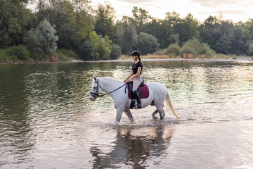 Girl riding a snow white horse down the calm river water with forest greenery reflections from the...