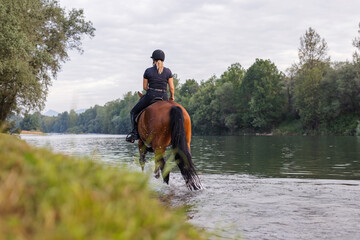 Female horseback rider in a black jockey outfit riding a chestnut horse along the river at sunset....
