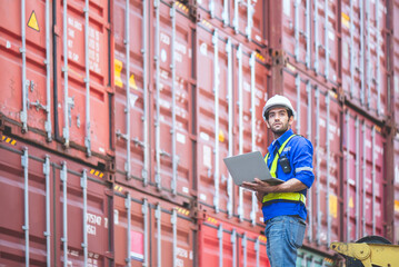 Engineer or cargo worker wears PPE standing in front of cargo stacked container yard and holding laptop and looking at the sky. concept logistic delivery management for industrial.