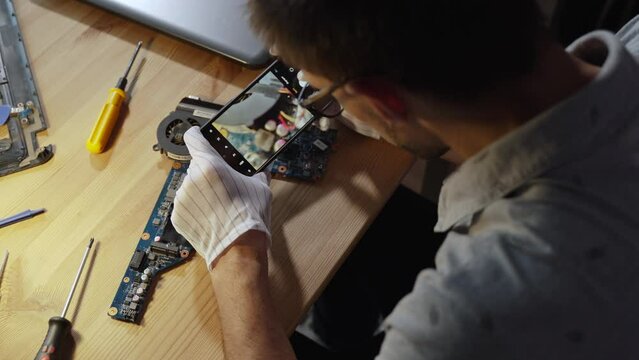Computer repairman photographs details of motherboard before repairing at workplace, over the shoulder. Technical engineer fixes with photo cause of breakdown, problematic electronic components.