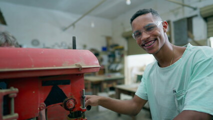 One happy young black carpentry apprentice using industrial machine, turns toward camera smiling. Friendly portrait of a worker engaged with job occupation