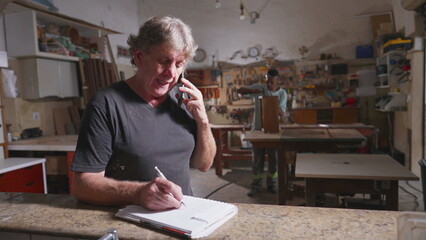 Small business owner of carpentry shop writing down orders by phone. Carpenter takes notes on paper while speaking with customer client, Apprentice and equipment in background