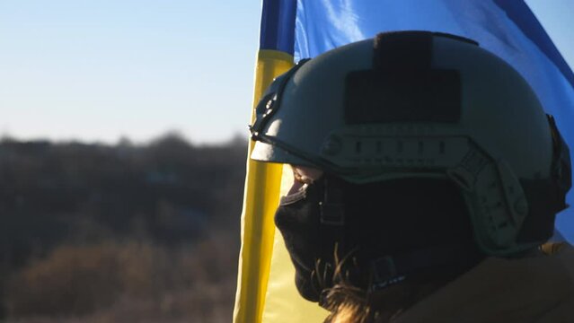 Dolly shot of girl in military helmet and balaclava holds a waving national flag. Female soldier of ukrainian army lifted blue-yellow banner at countryside. Invasion of territory Ukraine. Close up