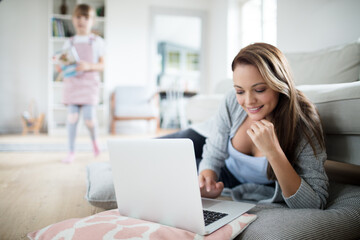 Young mother using a laptop in the living room with her daughter behind her