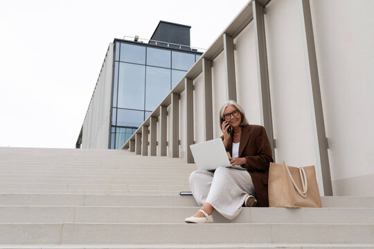 a gray-haired middle-aged woman with a bob hairstyle dressed in a jacket and trousers works remotely sitting on the steps with a laptop on the street