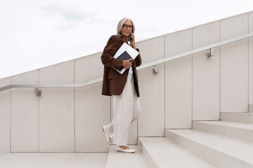 a gray-haired middle-aged woman with a bob hairstyle dressed in a jacket and trousers walks around the city against the backdrop of a business center and offices