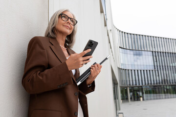 a well-groomed middle-aged woman in glasses dressed in a brown jacket stands leaning against the wall of a business center