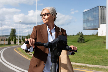 well-groomed middle-aged woman in glasses dressed in a brown jacket rides an electric scooter